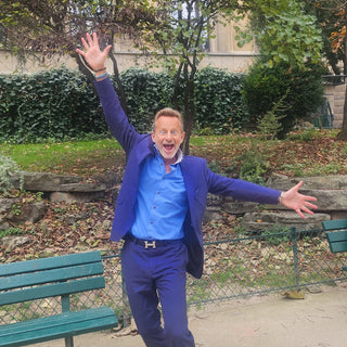 Geoffroy Gardair, co-fondateur de Dohrnii, boutique spécialisée en phycocyanine - il est habillé d'un costume bleu marine et d'une chemise bleu ciel et ouvre ses bras en grand pour exprimer sa joie de vivre - Il se tient dans un parc, l'on aperçoit un banc à côté de lui et des arbres en arrière plan